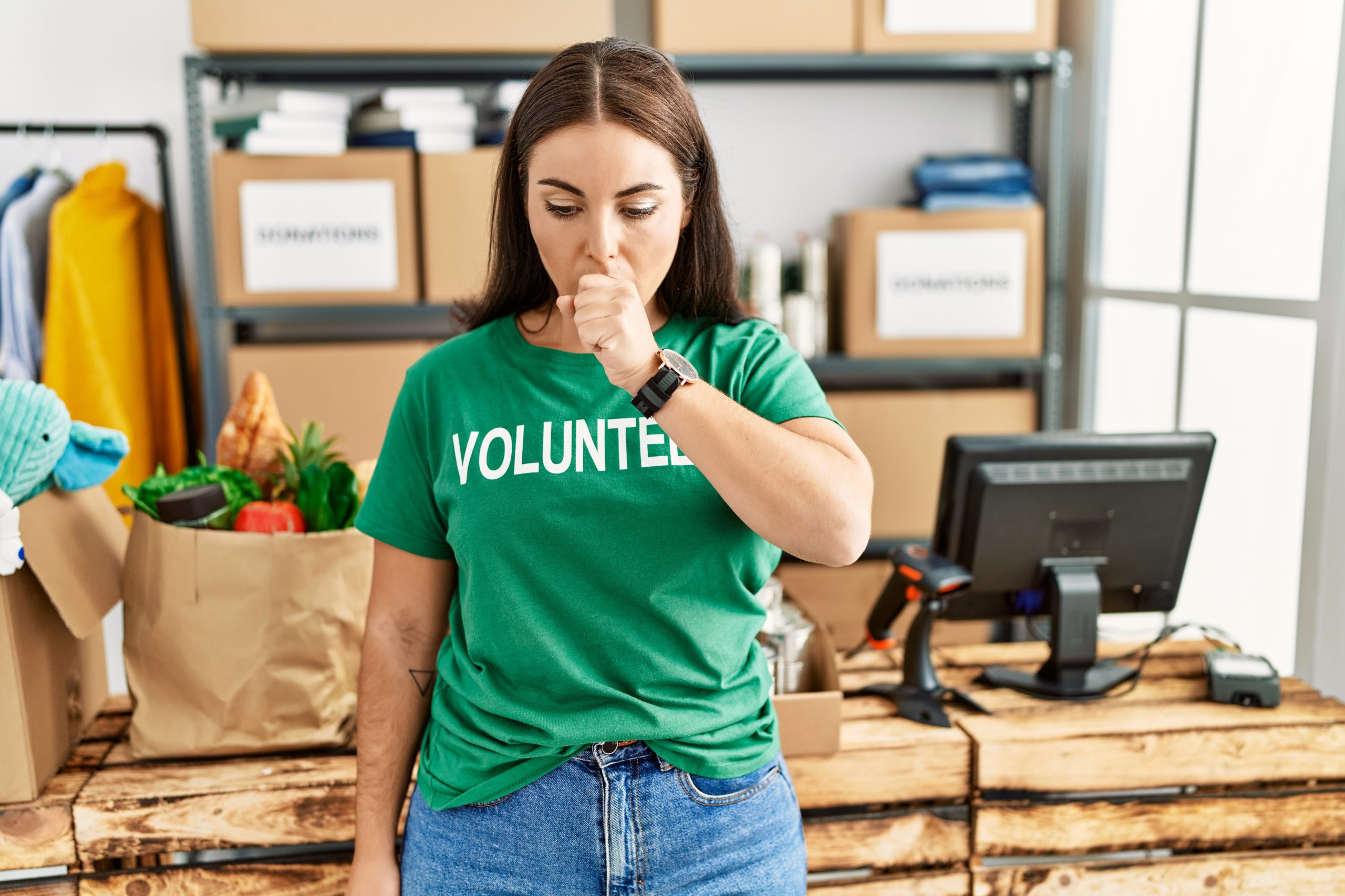 Young brunette woman wearing volunteer t shirt at donations stand feeling unwell and coughing as symptom for cold or bronchitis. health care concept.